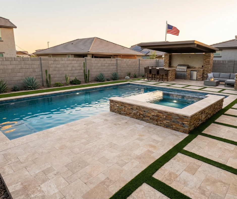 Modern Arizona backyard with a rectangular pool and raised stone-veneer spa, travertine paver deck with synthetic turf ribbon accents, a covered outdoor kitchen and bar area with stacked stone columns, an American flag, and desert landscaping with cactus and agave along the block wall.