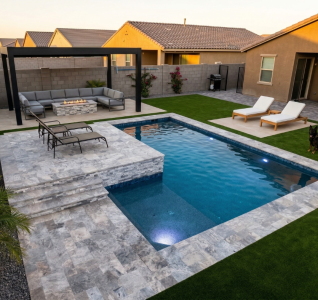 Elevated view of a contemporary geometric pool with silver travertine coping and raised deck, black metal pergola over a sectional lounge and stone fire pit, white chaise lounges on the paver patio, synthetic turf throughout, and a German Shepherd relaxing poolside at dusk.