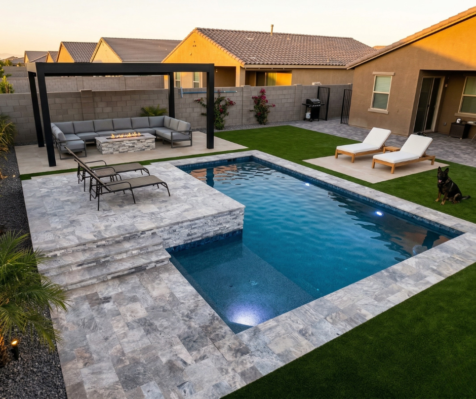 Elevated view of a contemporary geometric pool with silver travertine coping and raised deck, black metal pergola over a sectional lounge and stone fire pit, white chaise lounges on the paver patio, synthetic turf throughout, and a German Shepherd relaxing poolside at dusk.