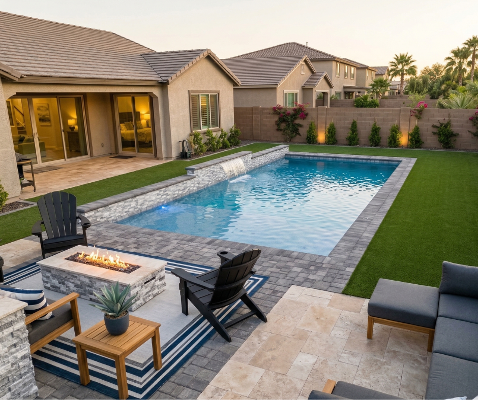 Arizona backyard at dusk with a rectangular pool featuring a raised stone water feature wall, gray paver coping, synthetic turf surround, a stone-veneer fire pit with black Adirondack chairs on a navy striped outdoor rug, a modern sectional sofa, and sliding glass doors opening to the home's interior.